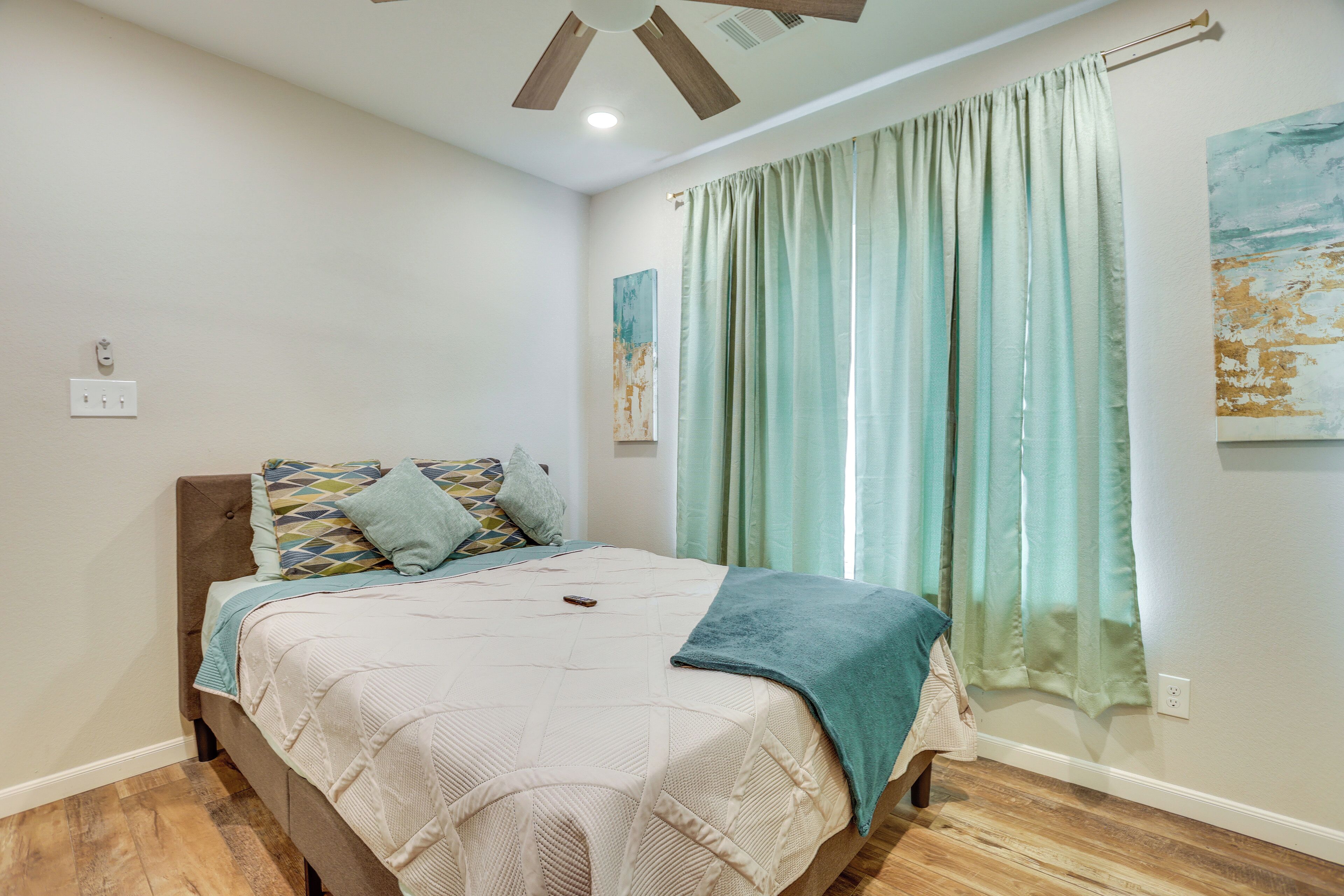 Un dormitorio con una cama, un ventilador de techo, una ventana con cortinas y una obra de arte colgada en la pared.