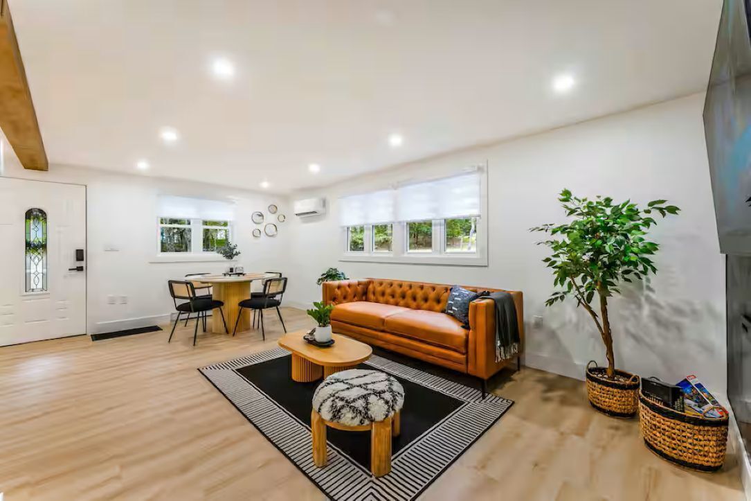 A modern living room with a sofa, coffee table, and chairs, a dining area with a table and chairs, and a large window with blinds.