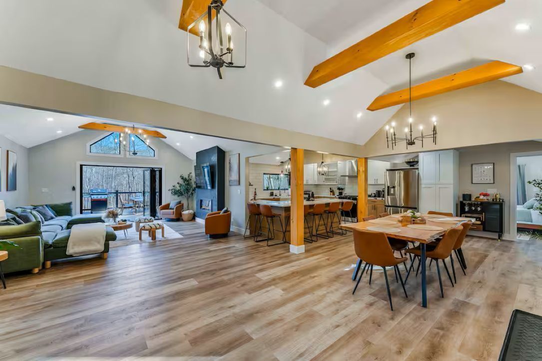 A spacious living area with a high ceiling, wooden beams, a dining table with chairs, and a kitchen in the background.