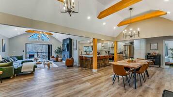 A spacious living area with a high ceiling, wooden beams, a dining table with chairs, and a kitchen in the background.