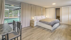 Un dormitorio con cama, una mesa de comedor con sillas, una mesita de noche con lámpara, una ventana con cortinas y vista a una piscina.