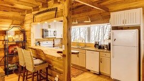 A wooden cabin interior with a kitchen area, bar stools, and a window with a view of trees.