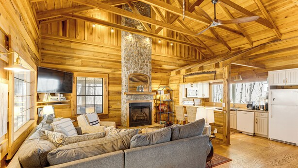 A log cabin interior with a fireplace, a large sofa, wooden walls, and a kitchen area.