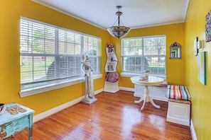 Una habitación con paredes amarillas, ventanas amplias con persianas, una mesa blanca sobre pedestal, un banco y una lámpara decorativa en el techo.