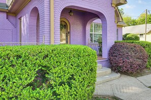 Una casa con fachada morada, una puerta principal y un pequeño jardín delantero con seto y arbusto.