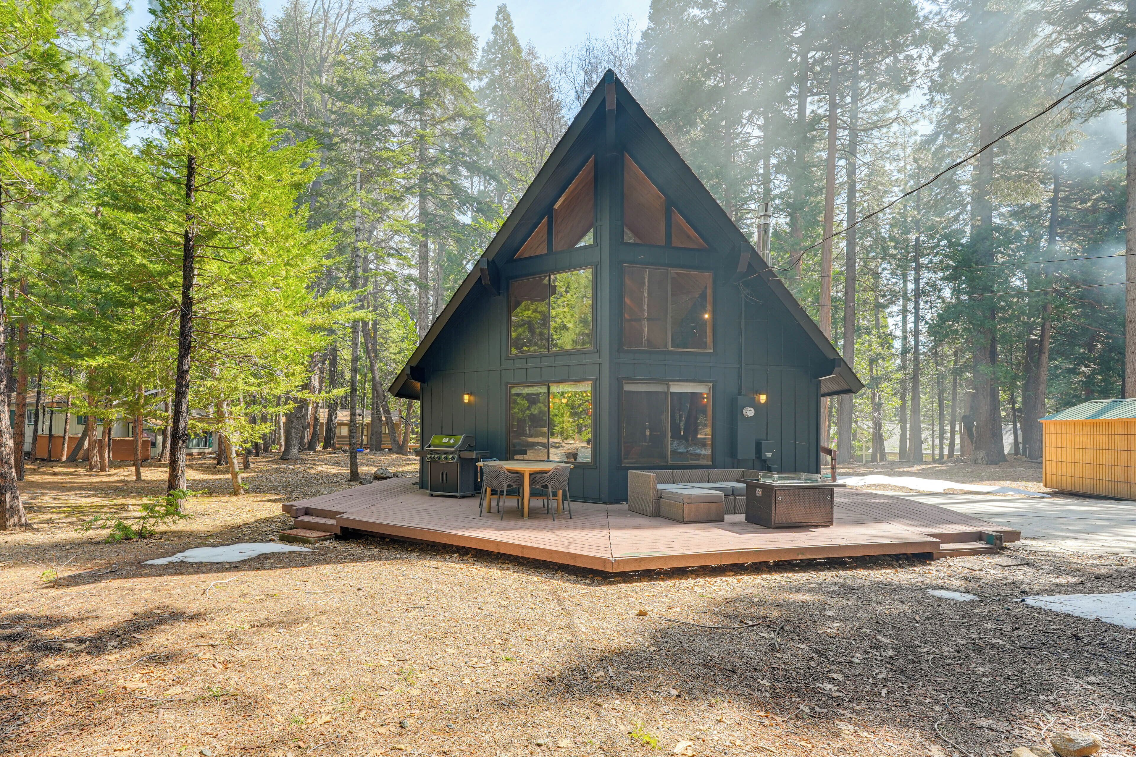 A cabin with a deck, outdoor seating, and a grill area surrounded by a forest.