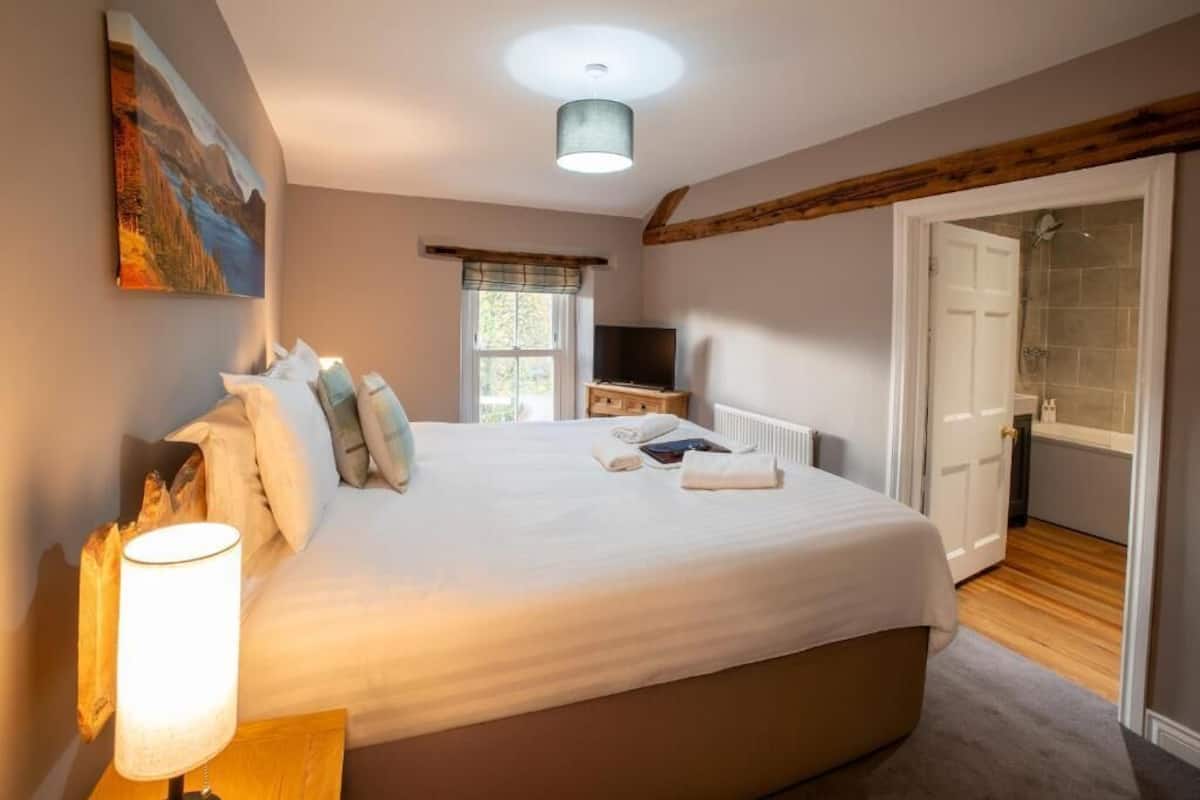 Un dormitorio con una cama, una lámpara de noche, un televisor, una ventana con cortinas y una puerta que da al baño.