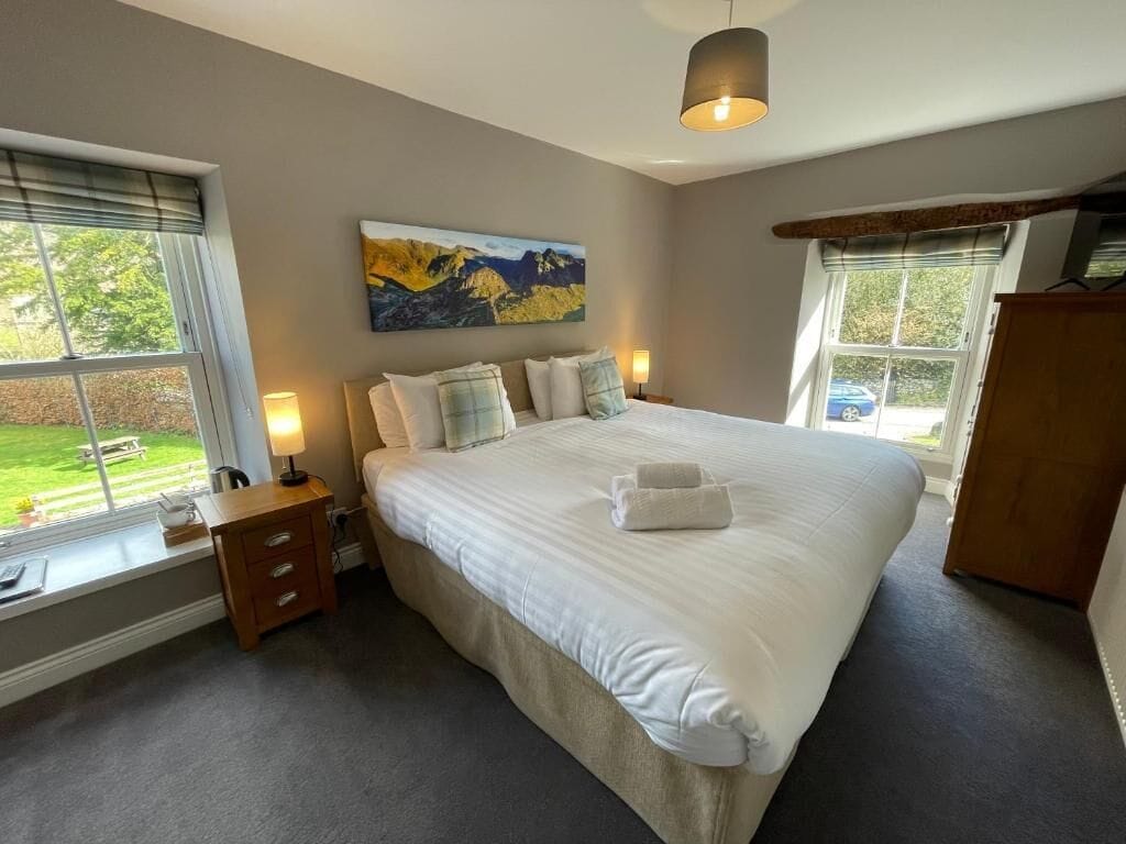 A bedroom with a large bed, a nightstand, a lamp, a painting on the wall, and a window with a view of the outdoors.