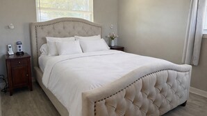 Un letto rifatto con un'elegante testiera imbottita, un comodino con un telefono e un piccolo elettrodomestico, e una finestra con persiane.