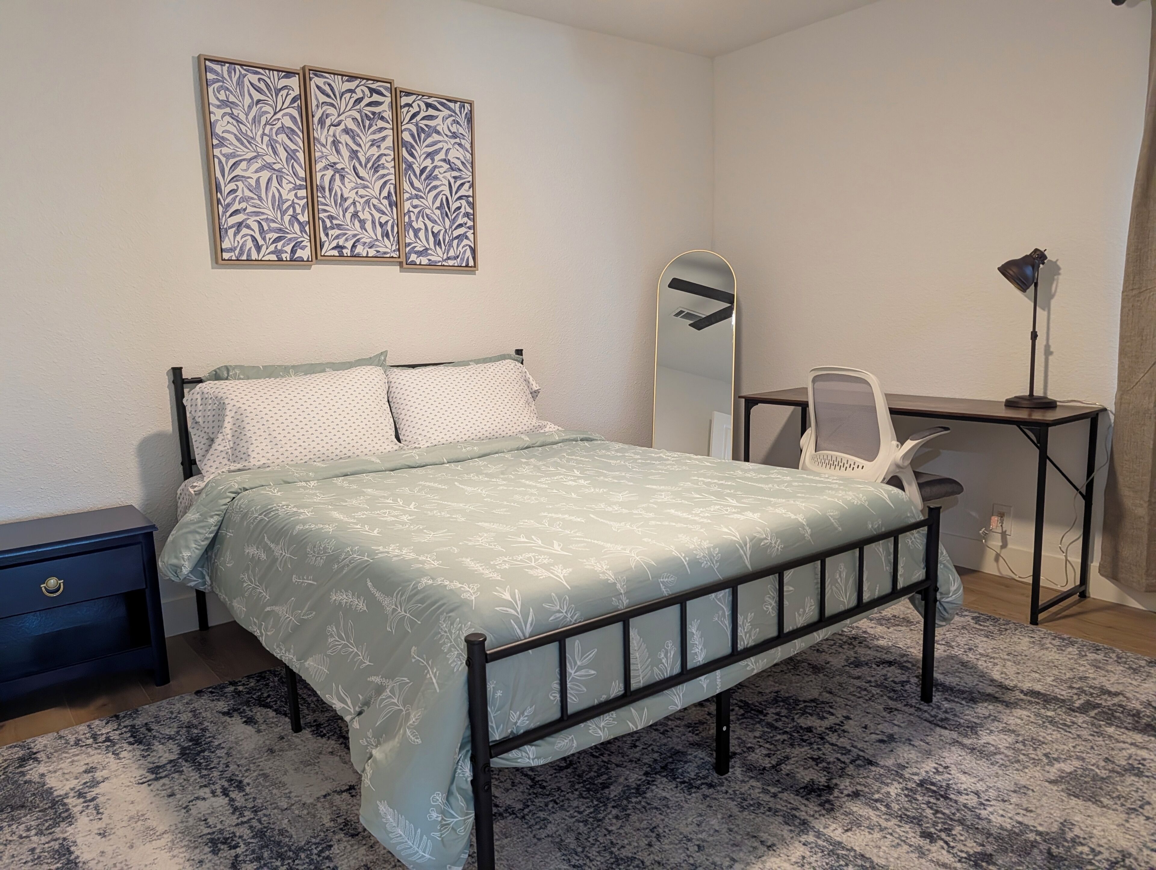 A bedroom with a bed, a desk, a chair, a nightstand, and a wall-mounted mirror.