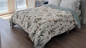 Een slaapkamer met een bed, een nachtkastje, een plafondventilator, een raam met jaloezieën en een aan de muur hangend schilderij.