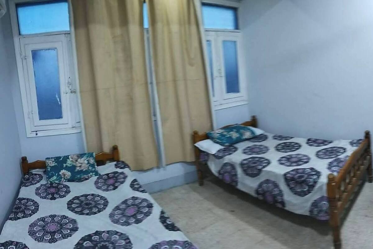 A bedroom with two single beds, a wooden wardrobe, a television mounted on the wall, and a small table with a patterned tablecloth.