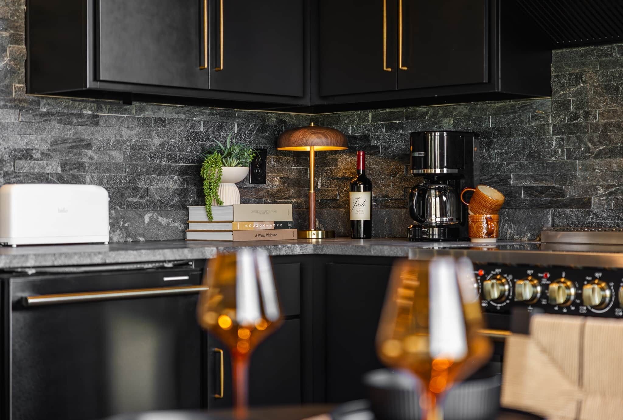 A modern kitchen with dark cabinetry, a stone backsplash, a coffee maker, a wine bottle, and a lamp.