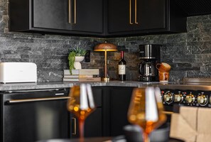 Eine moderne Küche mit dunklen Schränken, einer Steinwandverkleidung, einer Kaffeemaschine, einer Weinflasche und einer Lampe.