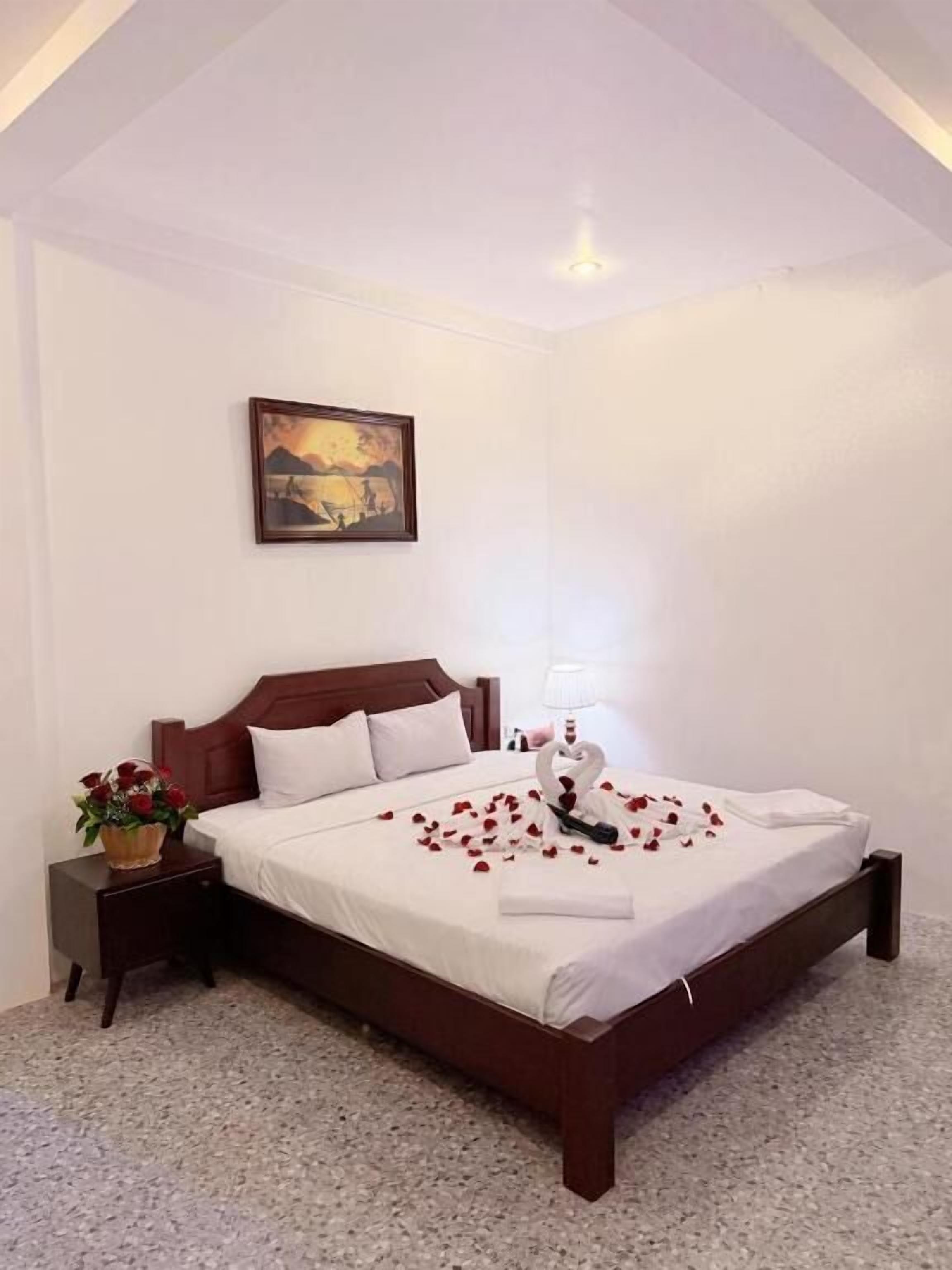 Una cama bien hecha con sábanas blancas, cabecera de madera, mesita de noche con un jarrón de flores y un cuadro en la pared.