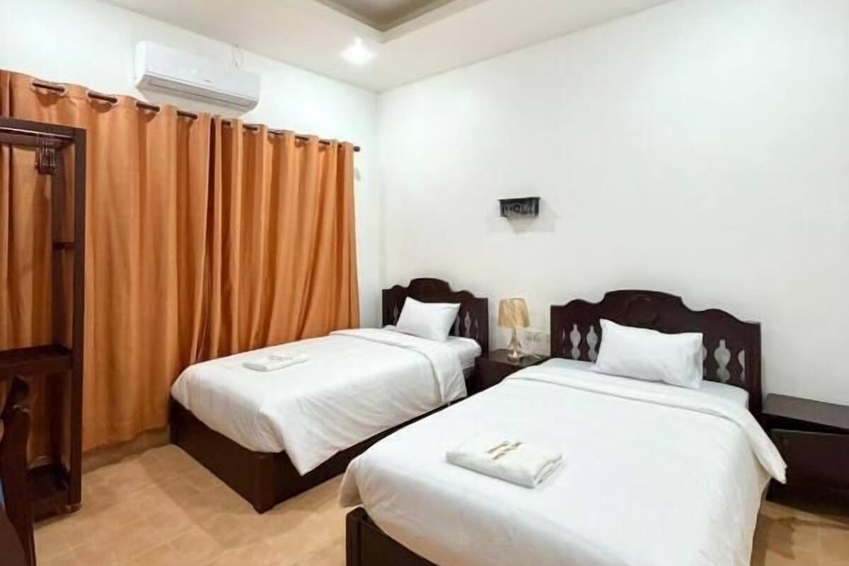 A hotel room with two single beds, a wooden desk, an air conditioning unit, and a window with orange curtains.