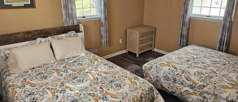 A bedroom with two beds, a wooden headboard, a small bedside table, a wall-mounted heater, and a framed picture.