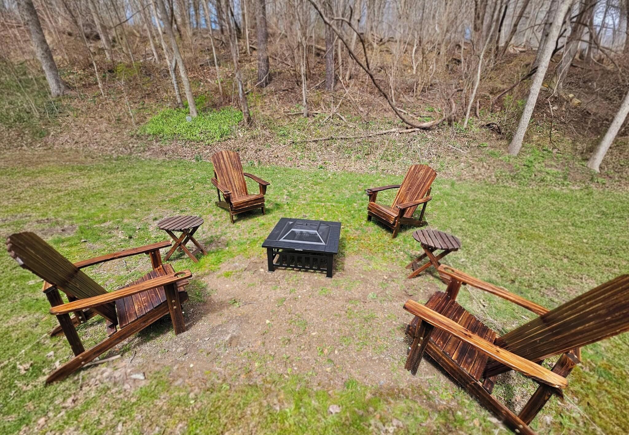 Quatre chaises Adirondack en bois sont disposées en cercle autour d’un foyer central, dans un espace extérieur verdoyant agrémenté d’arbres en arrière-plan.
