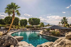 Ein Pool mit klarem Wasser, umgeben von Felsen und Palmen, mit Liegestühlen und Sonnenschirmen.