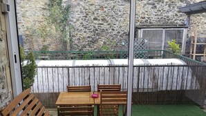 A wooden lounge chair and table set up near a sliding glass door, with a stone wall and greenery visible outside.