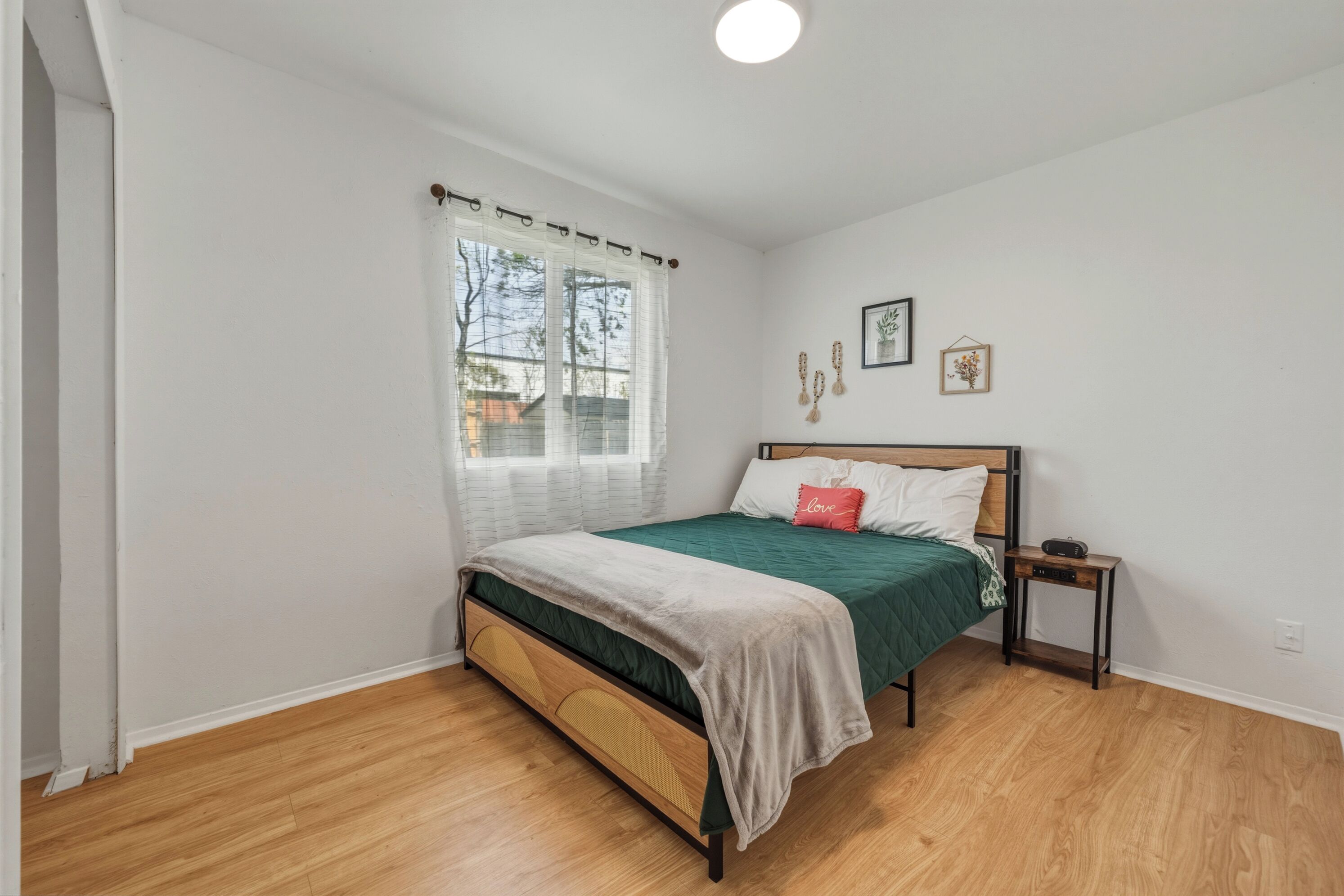 A bedroom with a bed, a nightstand, a window with curtains, and a wall with framed pictures.
