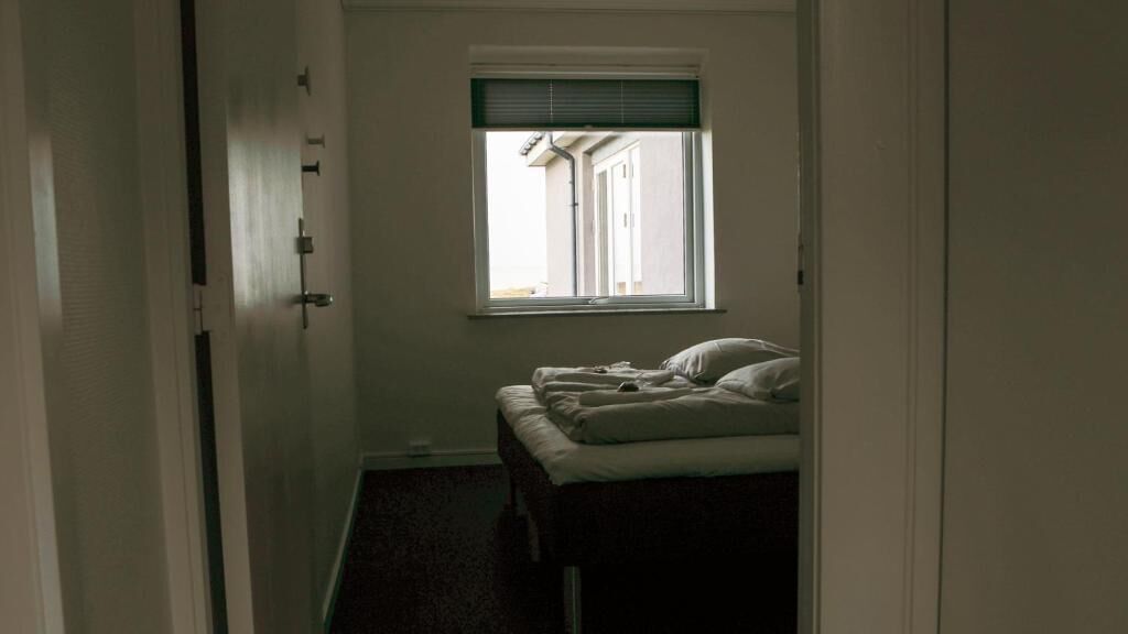 A narrow room with a bed, a window with blinds, and a door.