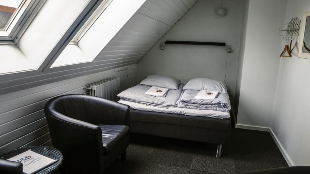 Quarto pequeno e pouco iluminado, com teto inclinado, uma cama simples com almofadas e cobertor, uma cadeira e uma pequena mesa com um cardápio.