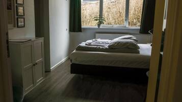 Ein Schlafzimmer mit Bett, Heizkörper, Fenster mit Blick ins Freie und einer Tür zu einem anderen Raum.