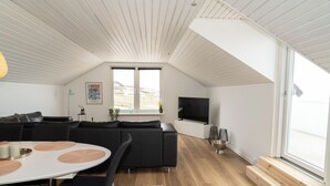 Un espacio habitable moderno estilo loft con techo alto, un ventanal grande, un televisor de pantalla plana, una zona de comedor con mesa redonda y un sofá modular negro.