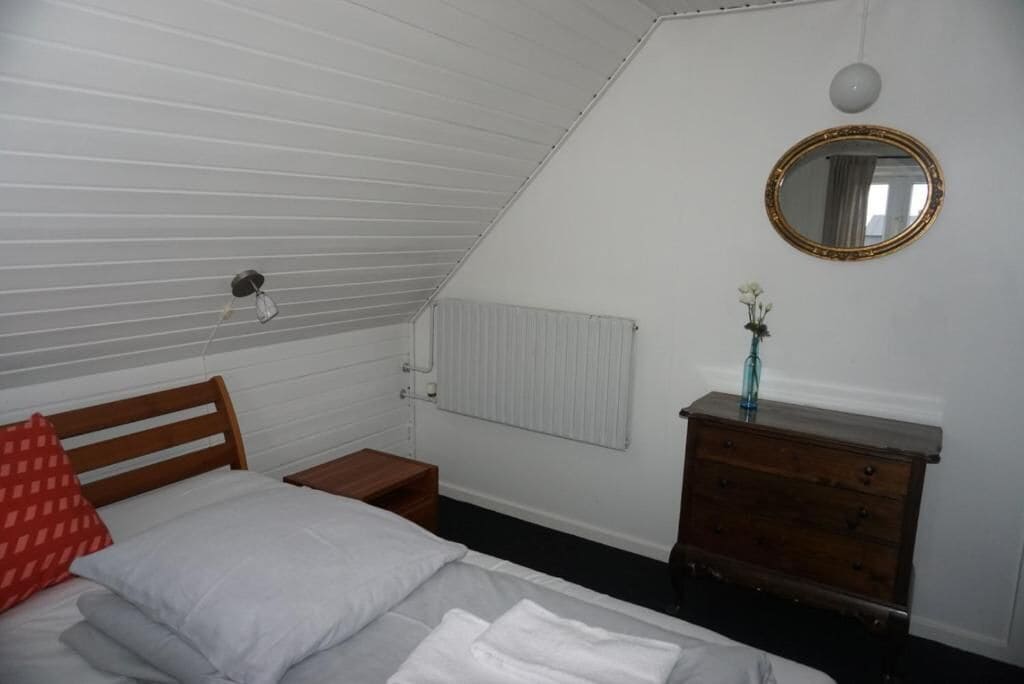 Un dormitorio con techo inclinado, una cama con almohadas, un aparador, un espejo y un radiador.