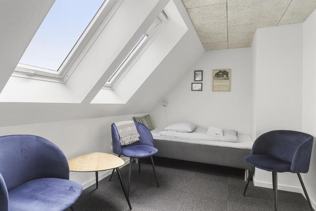 Una habitación con techo inclinado, tragaluces, dos sillas azules, una mesita redonda, un sofá con cojines y dos cuadros enmarcados en la pared.