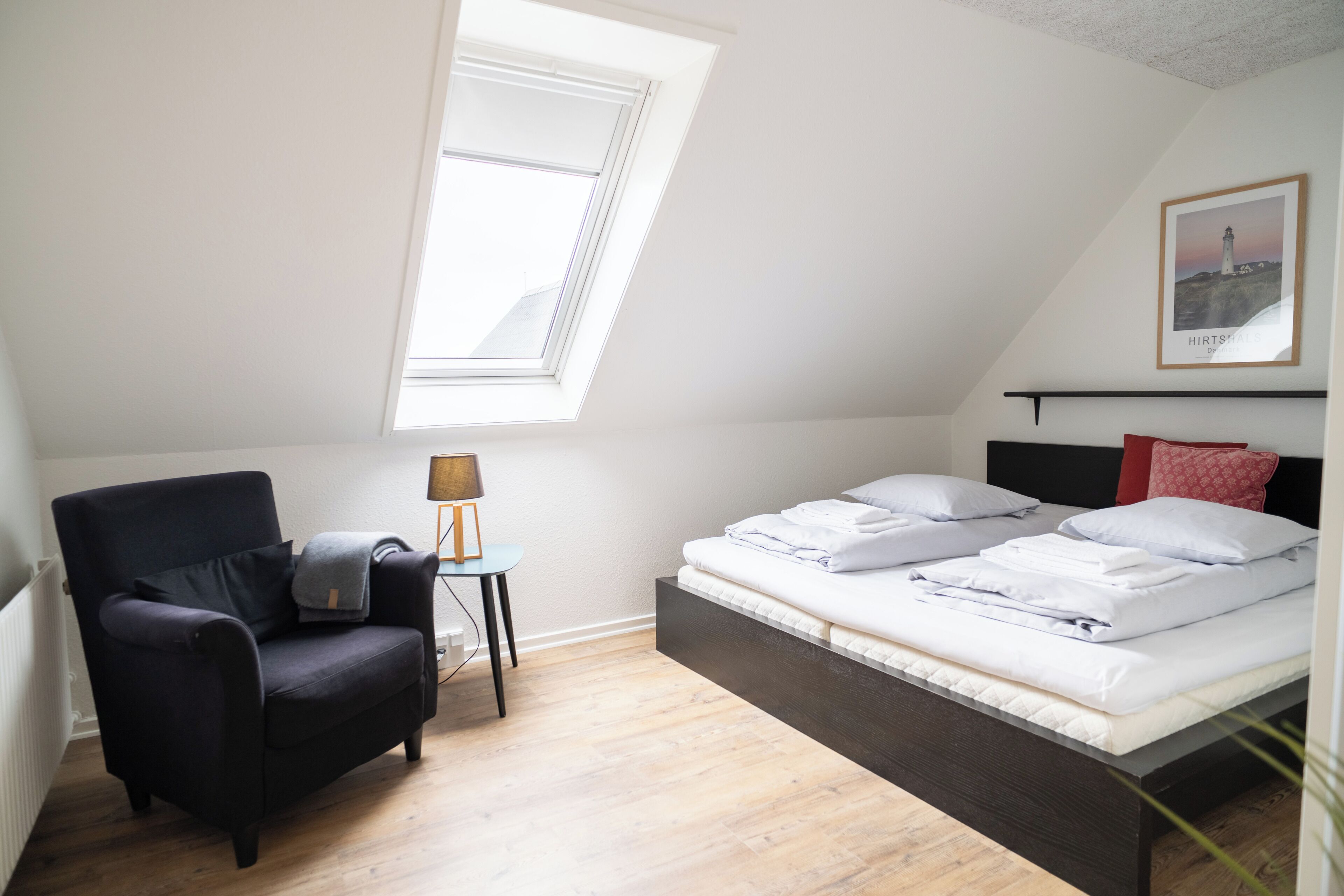 A room with a bed, a chair, a small table, a lamp, and a picture on the wall.