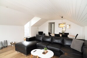 A modern living room with a black leather sectional sofa, a white coffee table, and a dining area with a table and chairs. - B&B by the Sea Hirtshals (Hirtshals)