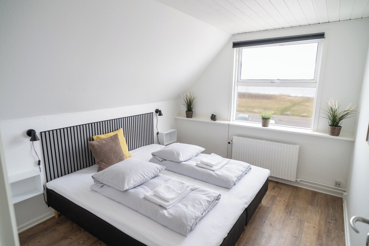 A bedroom with a bed, a radiator, a window with a view, and a shelf with plants.
