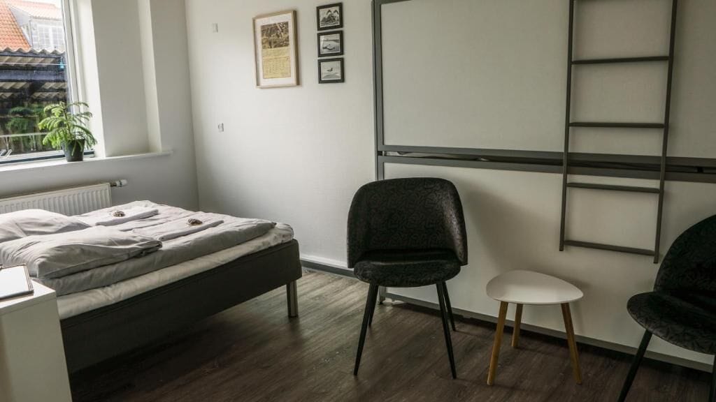 A bedroom with a bed, a chair, a small table, a radiator, a window with a view, and a wall-mounted shelf.