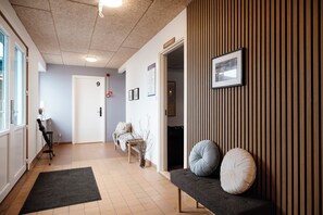 Un hall d’hôtel comprenant un comptoir d’accueil, un espace détente avec un banc et des coussins, une porte portant le numéro 9 et un panneau mural indiquant les numéros de chambre.