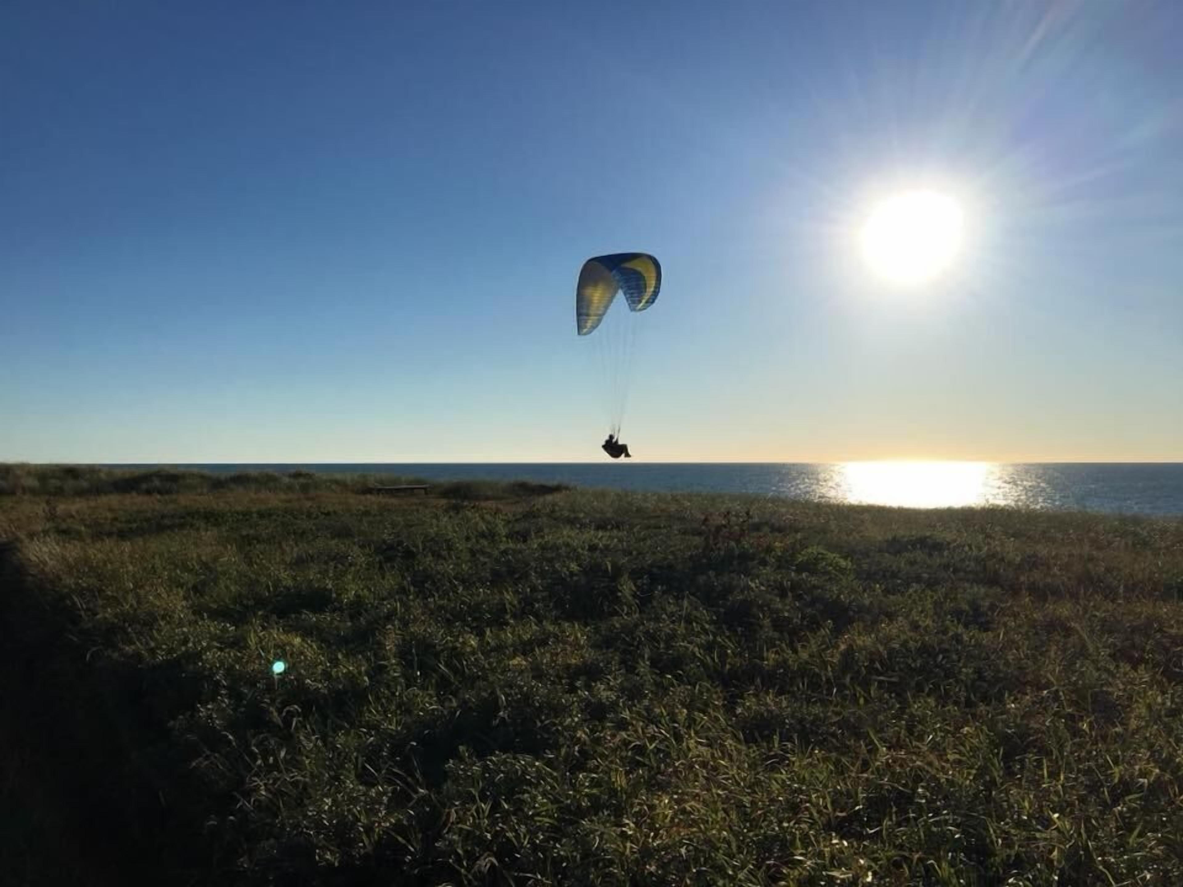Una persona practicando parapente sobre un campo cubierto de hierba, con una puesta de sol al fondo.