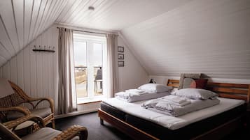 Una camera da letto con soffitto spiovente, un letto con lenzuola bianche, una sedia in vimini e vista panoramica tramite una porta vetrata.
