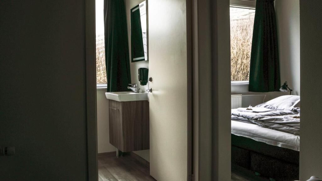 Un dormitorio con cama, ventana con cortinas, mesita de noche y baño con lavamanos y espejo.