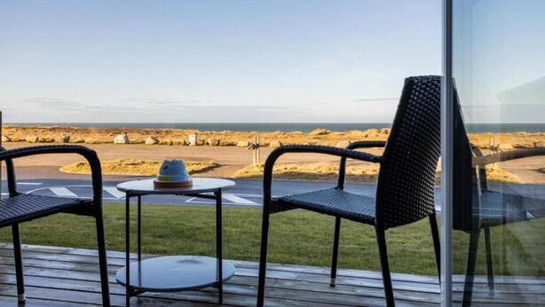 Terraza con dos sillas y una mesita pequeña, con vista a una playa rocosa y cielo despejado.