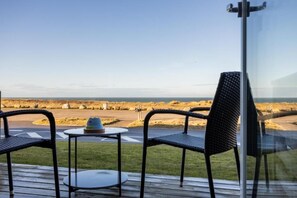 Terraza con dos sillas y una mesita pequeña, con vista a una playa rocosa y cielo despejado.