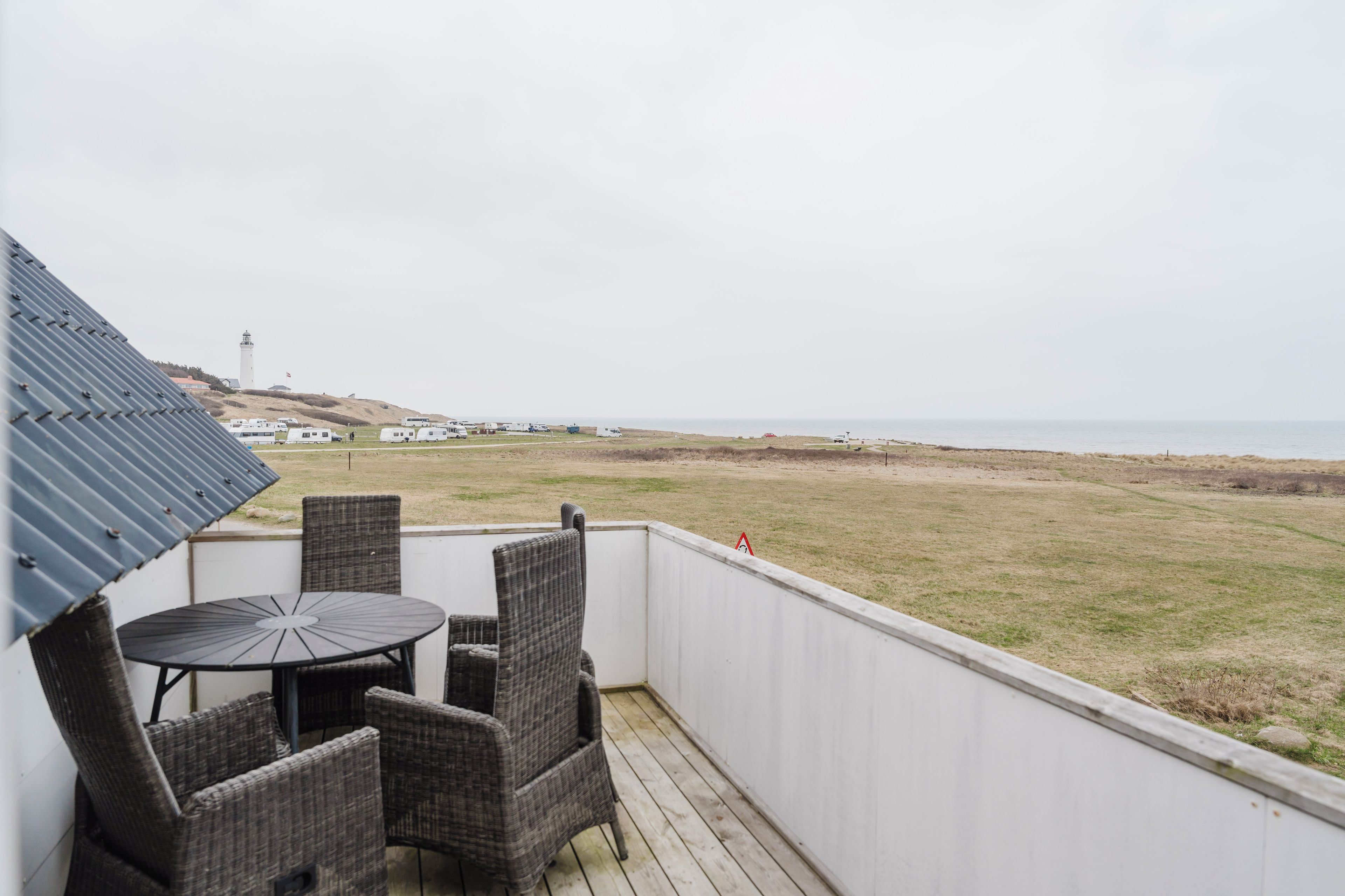 A balcony with wicker chairs and a table, overlooking a beach with a lighthouse in the distance.