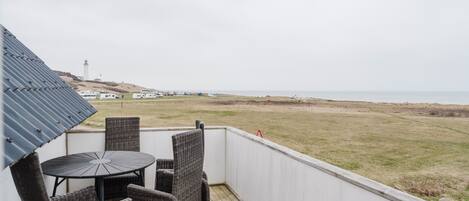 Un balcón con sillas de mimbre y una mesa, con vista a una playa y un faro a lo lejos.
