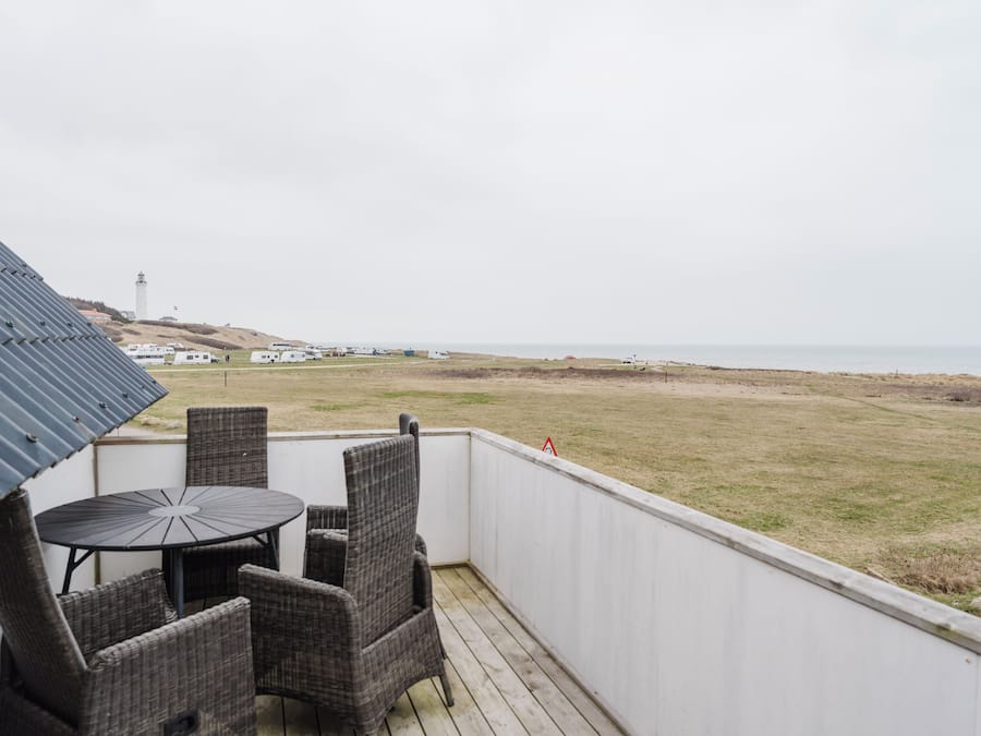 A balcony with wicker chairs and a table, overlooking a beach with a lighthouse in the distance.