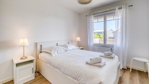 Un dormitorio con una cama, dos lámparas de noche, un ventanal con cortinas y una mesita blanca.