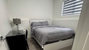 A bedroom with a bed, a desk, a chair, a lamp, and a window with blinds. - Stylish, conducive, and serene 2-bedroom suite located in the heart of Brighton, (Saskatoon)