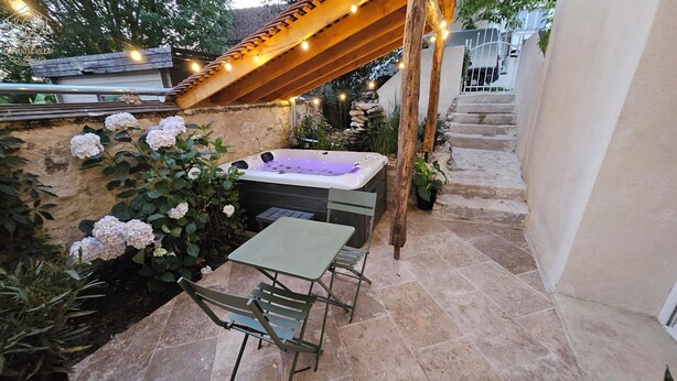 A patio area with a hot tub, a table, and chairs, surrounded by plants and a pergola with string lights. - THE FRENE SUITE (Montgé-en-Goële)