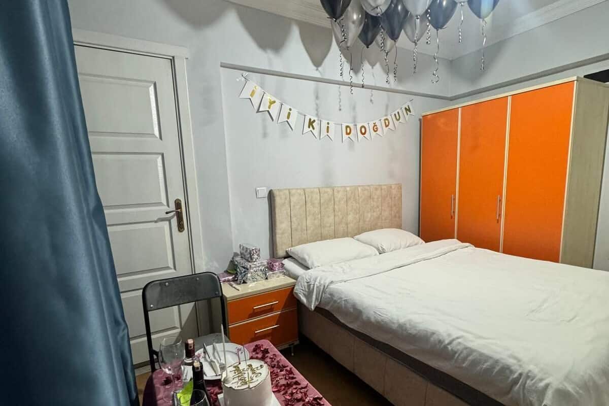 A bedroom with a bed, a nightstand, a table with a cake and glasses, a ceiling fan, and a banner on the wall.