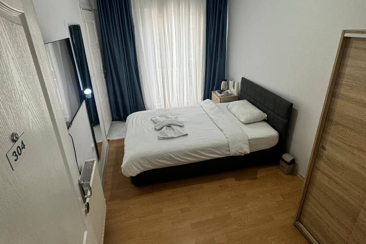 A bedroom with a bed, a bedside table, a nightstand, a ceiling fan, a window with curtains, and a door with a number.