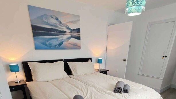 Ein Doppelbett mit weißem und dunkelbraunem Kopfteil, zwei Nachttischlampen, ein Gemälde einer Berglandschaft und eine Tür rechts.